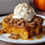 plate with a slice of pumpkin dump cake recipe with spice cake topped with whipped cream and a sprinkle of cinnamon. The dessert has a crumbly topping and a gooey, caramel-like filling.