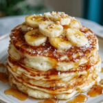 stack of banana pancakes recipe with sliced bananas on top, drizzled with syrup, and a sprinkling of powdered sugar.