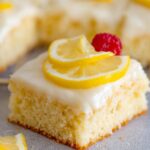 Close-up of a single square slice of lemon sheet cake recipe topped with white glaze, two thin lemon slices, and a raspberry on the side. The cake is moist and fluffy.
