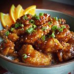 Close-up of crispy orange chicken recipe coated in a glossy orange sauce, garnished with sesame seeds and green onions, served over rice with slices of orange on the side.