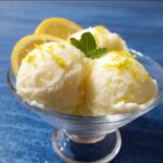 glass bowl with lemon sorbet recipe scoops garnished with fresh mint and lemon zest, alongside lemon wedges, set against a blue background