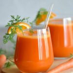 close-up of two glasses of fresh carrot juice garnished with a slice of orange and a sprig of green leaves. The juice has a vibrant orange color, and there are whole carrots in the background
