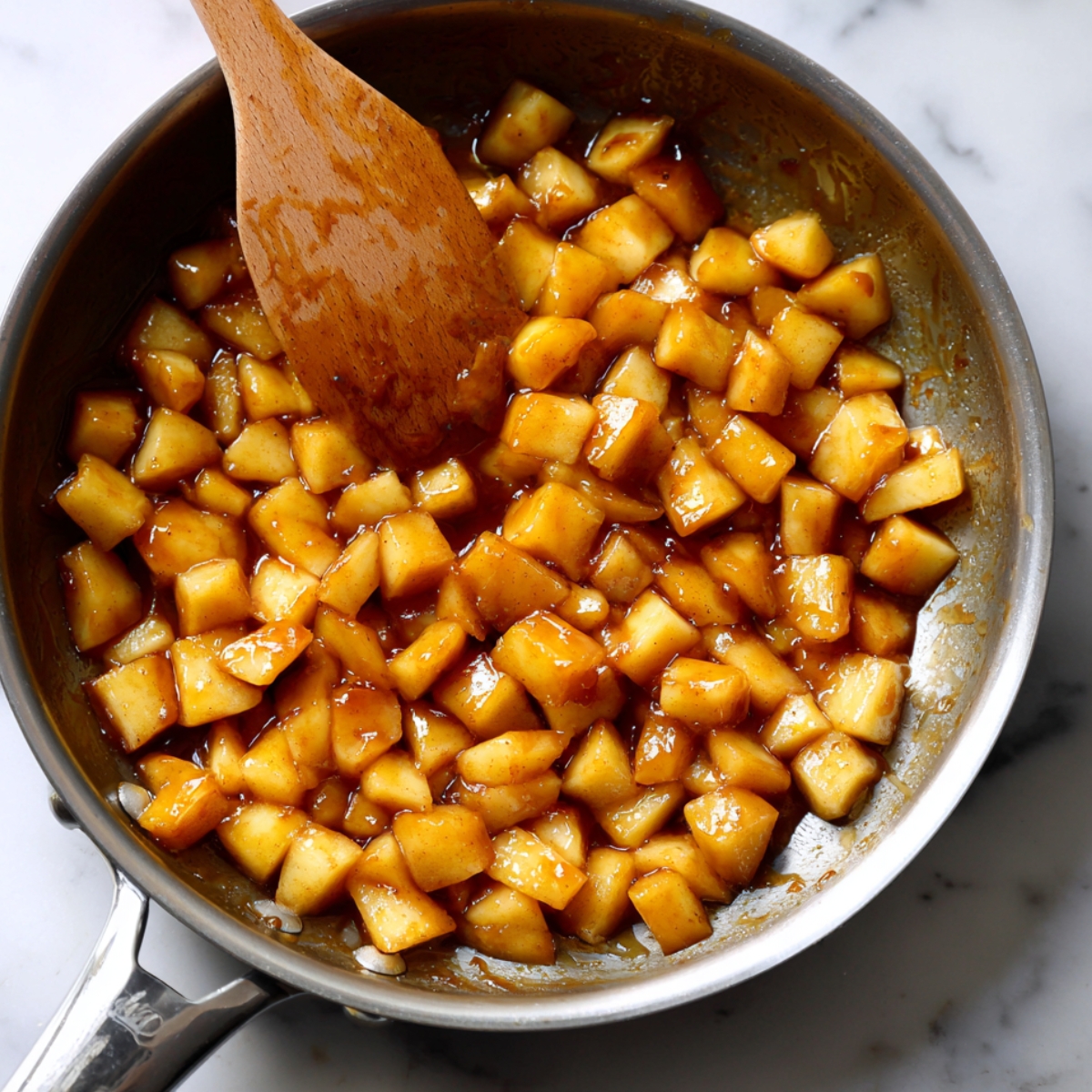 sauté pan with diced apples coated in a golden caramel sauce, with a wooden spoon resting in the pan. The apples appear soft and sweet, cooked in a syrupy sauce