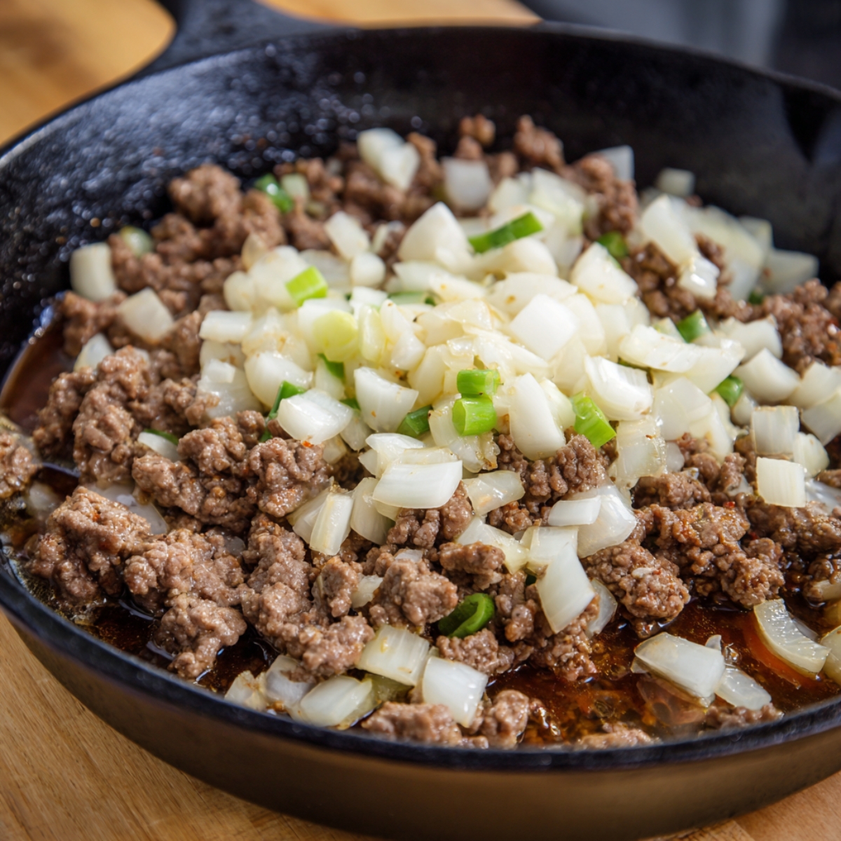 cast-iron skillet with ground beef being cooked, with chopped onions and green onions added, showing a sizzling cooking process.