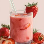 glass of a smooth pink strawberry juice recipe drink, garnished with whole strawberries. The drink looks cold and refreshing, and additional strawberries are placed around the glass