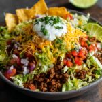 A colorful taco salad recipe with seasoned ground beef, shredded lettuce, diced tomatoes, onions, shredded cheese, sour cream, tortilla chips, and cilantro