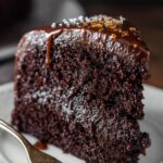 A close-up of a slice of rich, moistchocolate cake recipe topped with glossy chocolate ganache and a sprinkle of sea salt, placed on a white plate with a fork resting nearby.