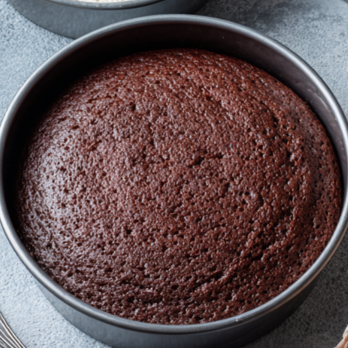 A freshly baked round chocolate cake in a metal pan, showing a perfectly even, risen surface, set against a light-colored background.