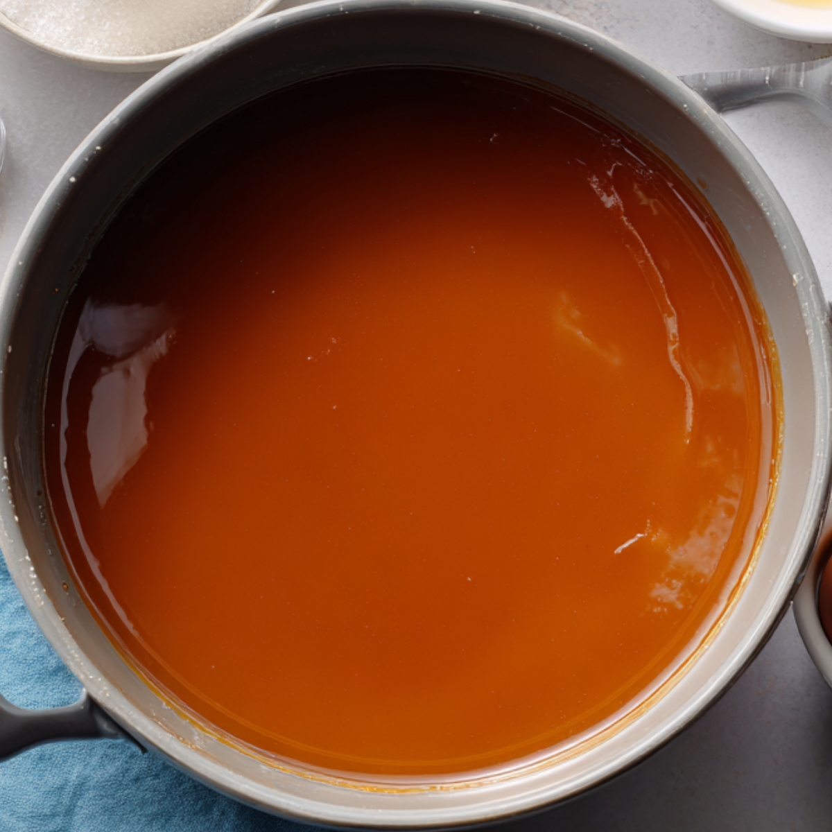 A saucepan filled with glossy, rich caramel sauce, freshly cooked and still warm, sitting on a blue kitchen towel with ingredients nearby.