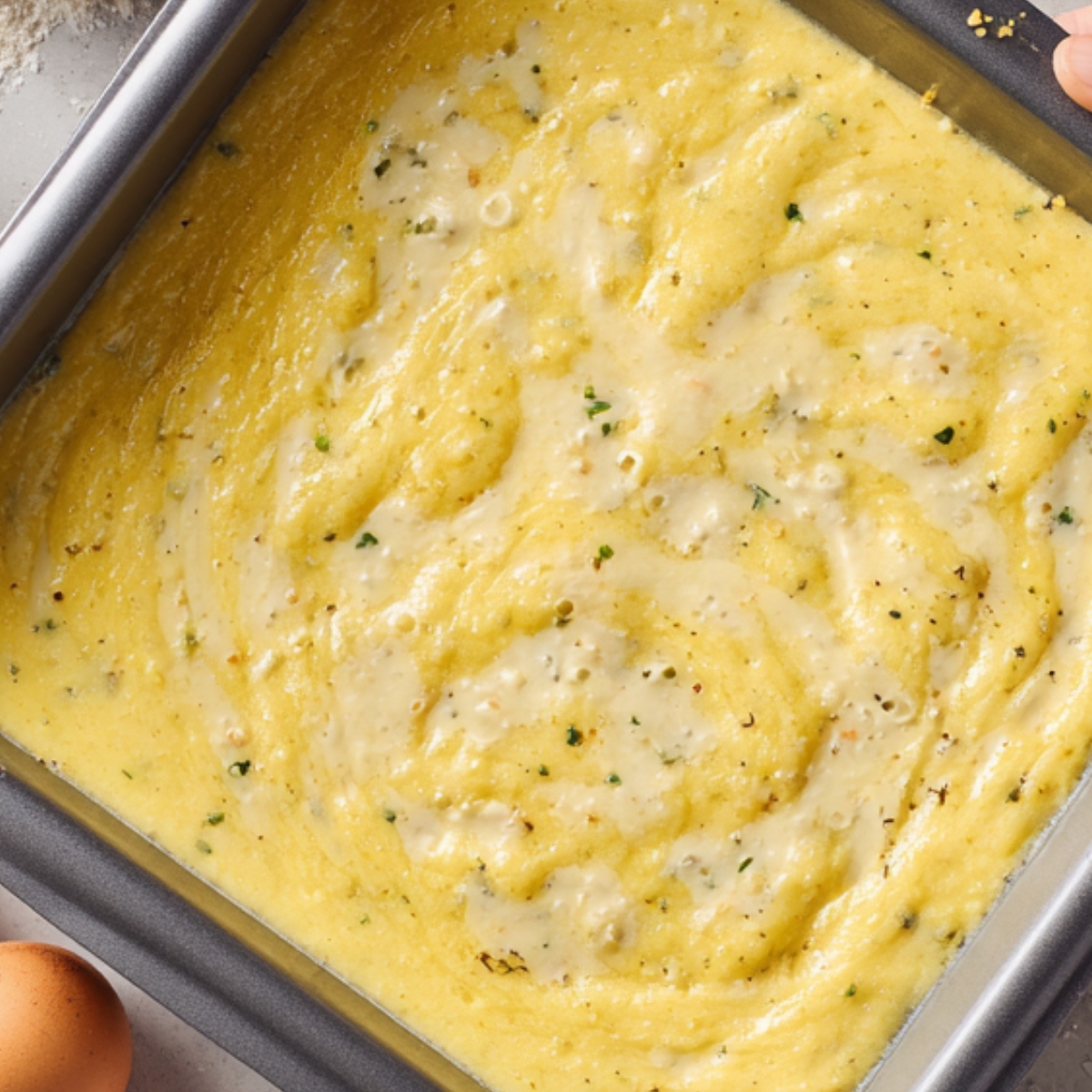 The image shows a close-up of a baking pan filled with a thick, yellow mixture — likely cornbread batter or a similar savory dish before baking. The batter appears smooth with small flecks of green herbs and possibly bits of pepper or seasoning scattered throughout. The texture is creamy and slightly swirled on the surface, suggesting it has just been poured or spread evenly into the pan. In the corner, part of an egg is visible, hinting at ingredients used in the mixture.