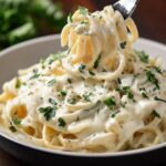 A close-up of a bowl of alfredo sauce recipe, fettuccine Alfredo, garnished with chopped parsley and cracked black pepper. A fork is twirling a portion of the pasta, showing its rich, velvety sauce.