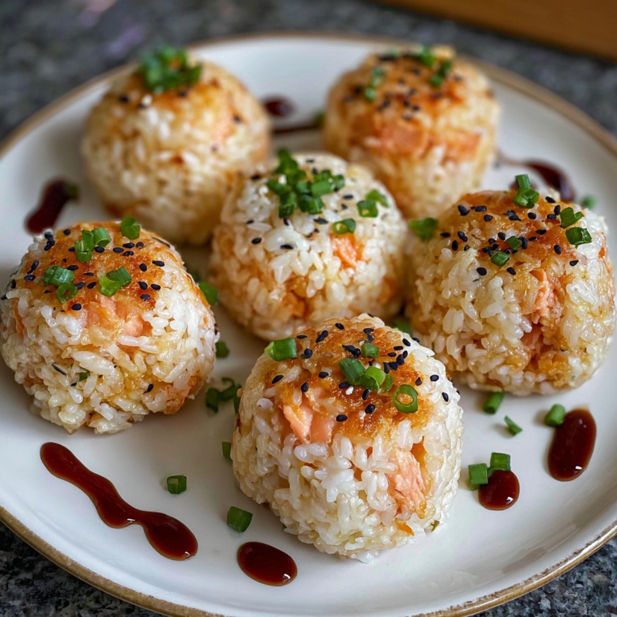 A plate of salmon rice balls , each containing pieces of salmon, garnished with chopped green onions, black sesame seeds, and drizzled with a soy-based sauce.