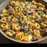 Close-up of creamy Philly philly cheese steak tortellini in a skillet — cheese-covered tortellini mixed with tender steak pieces, colorful peppers, and melted cheese garnished with chopped green onions.