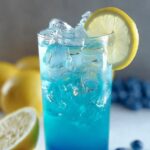 A glass of icy blue raspberry lemonade garnished with a lemon slice, with lemons and blueberries in the background.