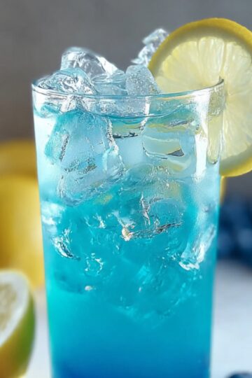A glass of icy blue raspberry lemonade garnished with a lemon slice, with lemons and blueberries in the background.
