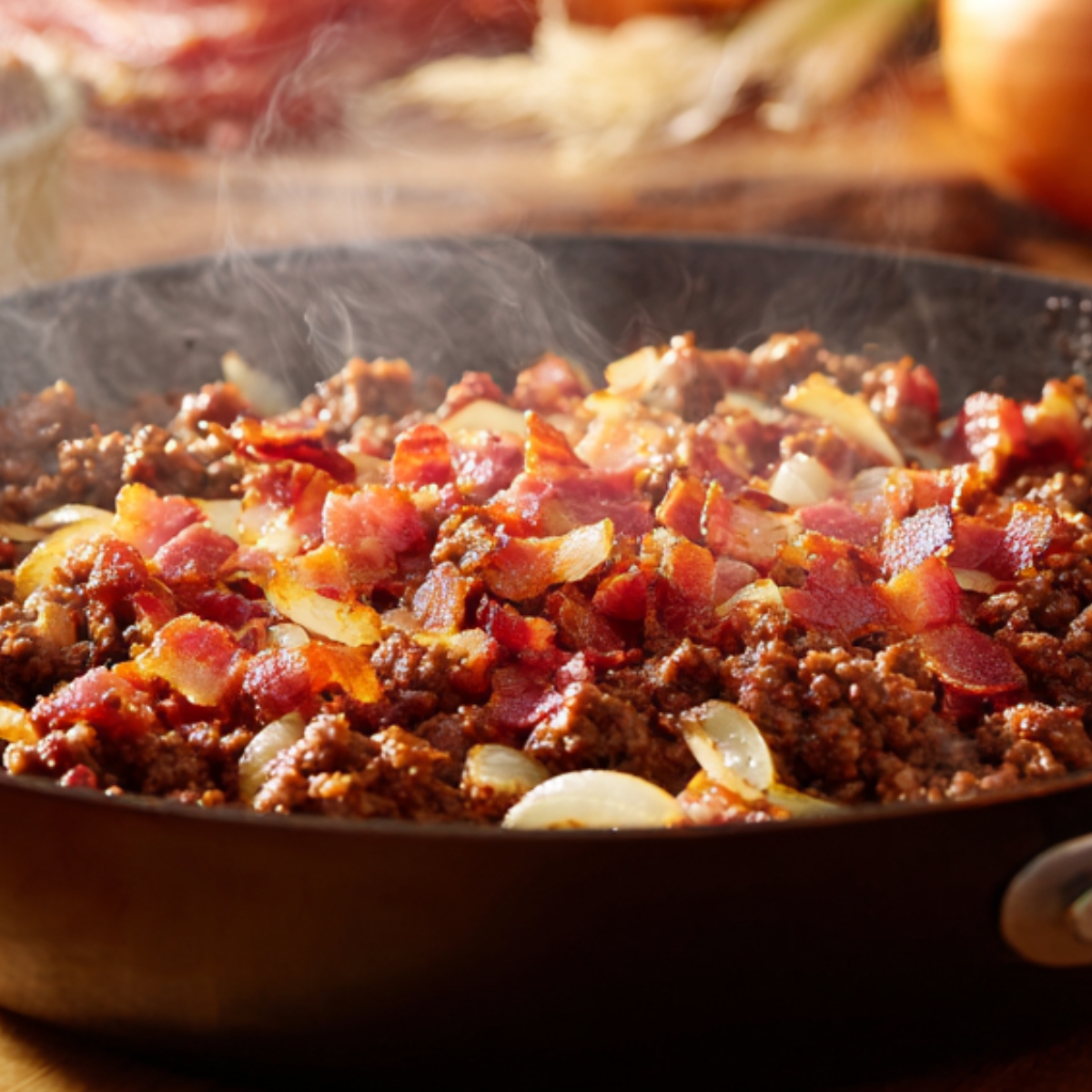A skillet filled with sizzling cooked ground beef, crispy bacon, and sautéed onions, steaming and golden brown.