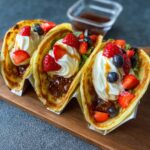 Three folded crepes served taco-style, filled with pancake tacos fresh strawberries, blueberries, and drizzled with caramel sauce, placed on a wooden board.