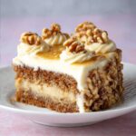 Slice of banana walnut cake layered with creamy frosting, decorated with whole walnuts and caramel drizzle on top, served on a white plate.