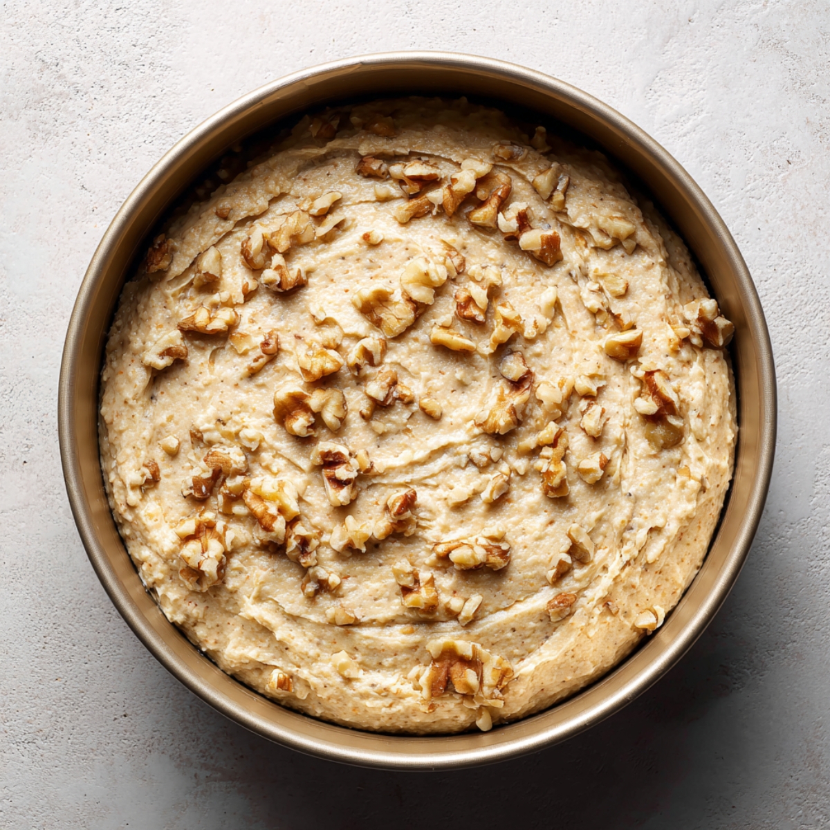 Round cake pan filled with banana walnut cake batter, topped with chopped walnuts before baking, on a light stone surface.