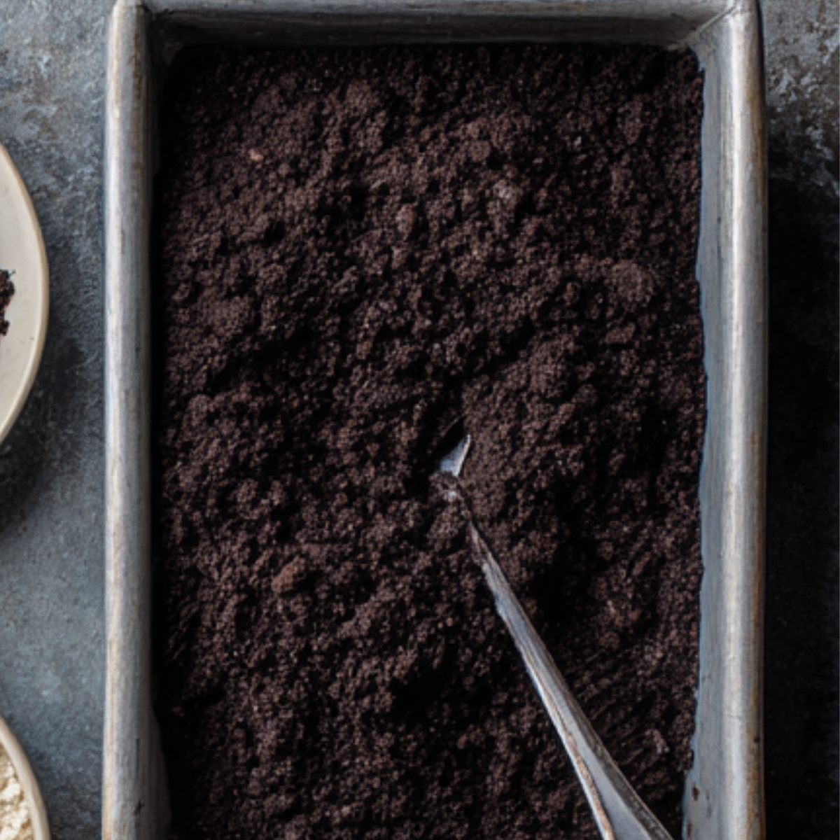 Close-up of crushed Oreo cookie crumbs in a metal baking pan with a spoon, ready to be used for a crust or topping.