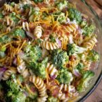 Creamy broccoli pasta salad mixed with rotini, cheddar cheese, red onions, and dressing in a large glass bowl.
