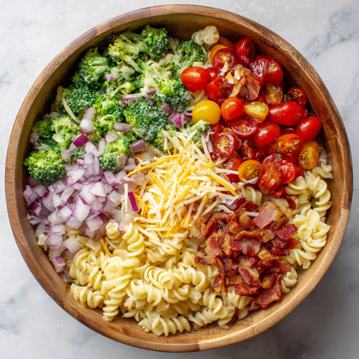 Wooden bowl filled with cooked rotini pasta, broccoli, cherry tomatoes, red onion, bacon, and shredded cheese arranged for salad preparation.