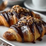 Two golden-brown chocolate croissant topped with drizzled melted chocolate and chopped nuts, dusted lightly with powdered sugar. They look glossy, flaky, and freshly baked, served on a white plate — a perfect presentation for pain au chocolat or chocolate almond croissants.