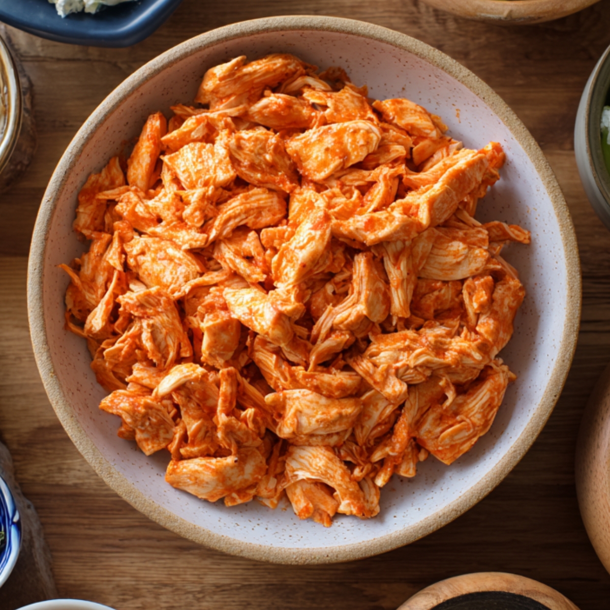 Bowl filled with shredded buffalo chicken coated in spicy orange sauce, placed on a wooden table with other ingredients around.