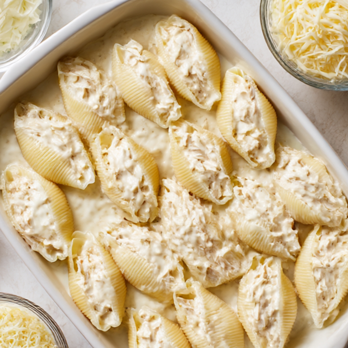 Unbaked chicken Alfredo stuffed shells arranged in a casserole dish with creamy white sauce and shredded cheese on the side.