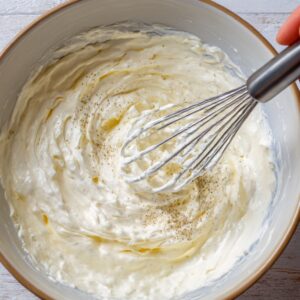 A close-up of a creamy dressing being whisked in a bowl, showing a smooth texture with some cracked pepper and a drizzle of oil on top.
