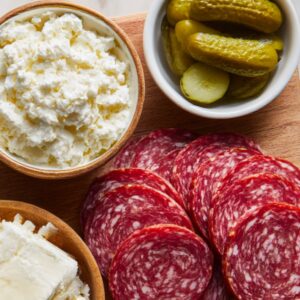 A wooden board with slices of salami, bowls of crumbled white cheese, feta, and pickles arranged for assembling appetizers.
