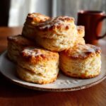 A plate stacked with golden brown, freshly biscuit recipe . They look flaky, buttery, and slightly dusted with flour, catching warm morning light beside a coffee mug.