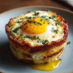 A single golden-brown baked eggs napoleon on a gray plate, featuring a perfectly cooked sunny-side-up egg on top, garnished with chopped herbs. The layers of pastry are visible and a bit of yolk has started to run out.