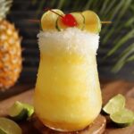 Frozen HOMEMADE pina colada punch in a glass with crushed ice, garnished with lime slices and a cherry, set on a wooden surface.