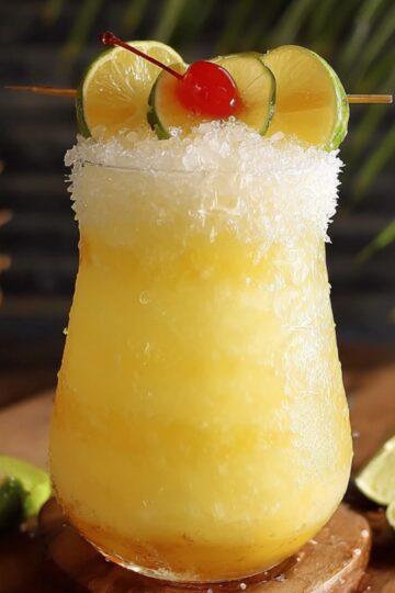Frozen HOMEMADE pina colada punch in a glass with crushed ice, garnished with lime slices and a cherry, set on a wooden surface.