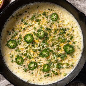 Cornbread batter topped with shredded cheese, jalapeño slices, and chopped chives in a cast-iron skillet before baking.