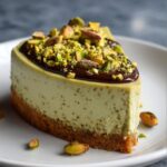 A slice of pistachio cheesecake with a nutty base and chocolate topping, garnished with pistachio pieces.