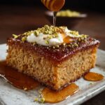 A thick square slice of Greek honey cake soaked in syrup, topped with whipped cream, chopped pistachios, and an extra drizzle of honey. The glossy surface and golden color highlight the cake’s moist, tender crumb.