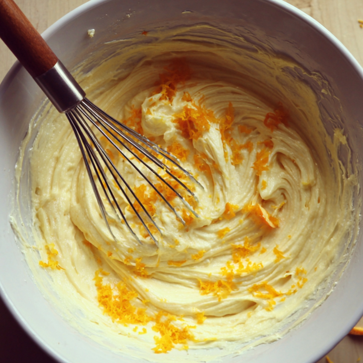 A bowl of creamy Greek honey cake batter with bright orange zest folded in, and a whisk resting inside. The smooth batter shows the blend of yogurt, olive oil, eggs, and citrus for a light, moist texture.