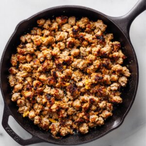 Close-up of browned crumbled breakfast sausage cooking in a cast iron skillet, with crispy golden edges and small bits of cooked seasoning throughout.