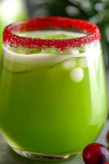 The Grinch punch recipe has a festive red sugar rim and is surrounded by fresh cranberries with blurred Christmas greenery in the background.