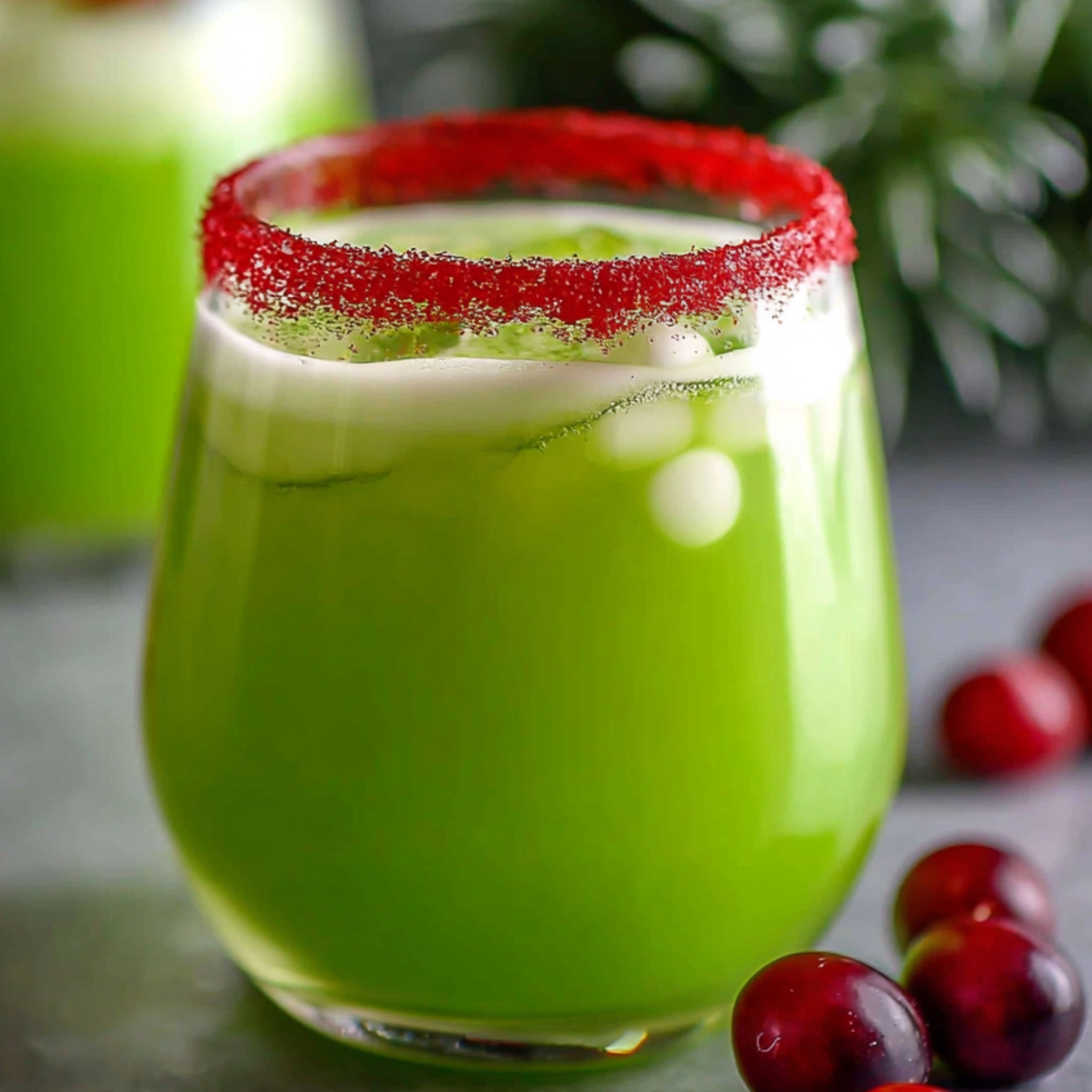 The Grinch punch recipe has a festive red sugar rim and is surrounded by fresh cranberries with blurred Christmas greenery in the background.