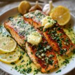 Two garlic butter salmon fillets on a white plate topped with garlic butter and herbs, surrounded by lemon slices and whole garlic cloves, with a buttery herb sauce.