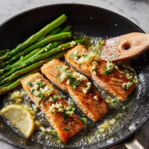 Three cooked salmon fillets in a skillet with asparagus spears, lemon wedge, garlic butter, and herbs on top.