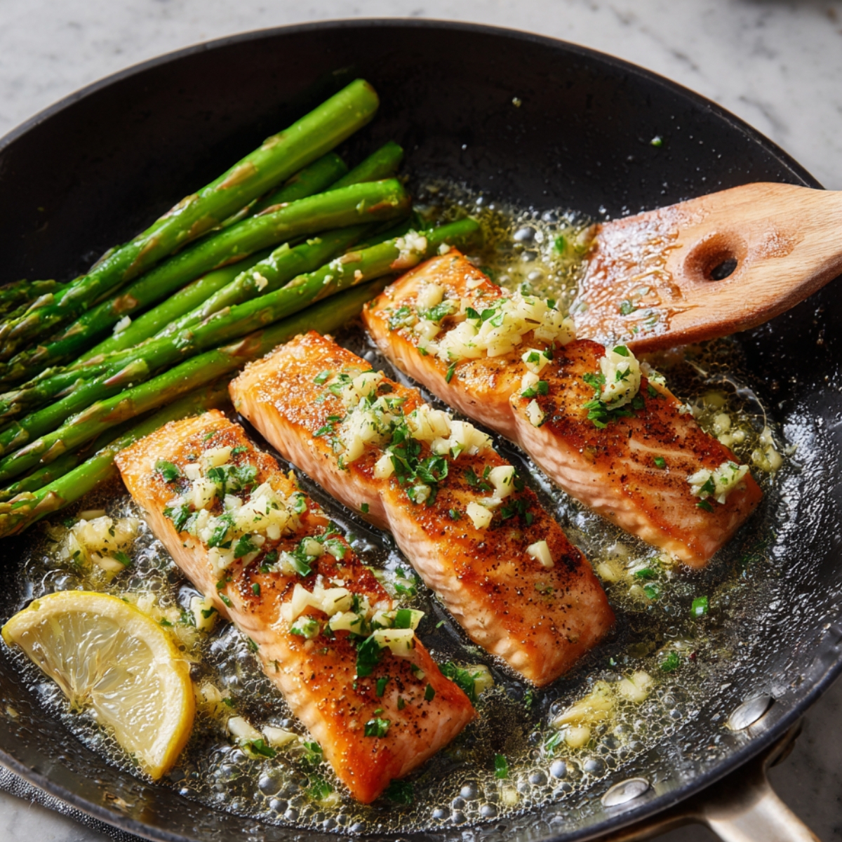 Three cooked salmon fillets in a skillet with asparagus spears, lemon wedge, garlic butter, and herbs on top.