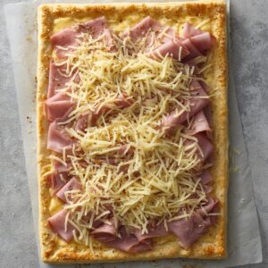Rectangular puff pastry tart topped with folded slices of ham and a generous layer of shredded cheese, sprinkled with black pepper, placed on parchment paper over a gray surface.
