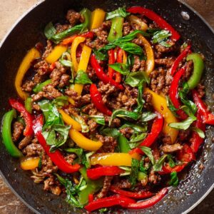 A colorful stir-fry dish in a black skillet featuring minced meat, red, yellow, and green bell peppers, and fresh basil leaves coated in a glossy sauce.