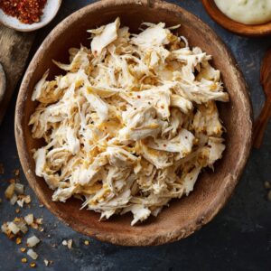 Wooden bowl of shredded cooked chicken seasoned lightly, ready to use as enchilada filling.