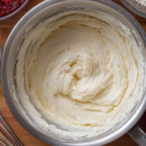 A bowl of classic vanilla buttercream frosting - pale cream color, thick and silky, with a whisk nearby. You can also see hints of strawberry puree or jam at the edge, likely ready to be added for flavor variations.