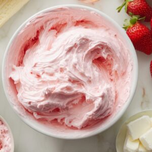 A bowl of fluffy pink strawberry buttercream frosting - light, whipped texture with a smooth swirl. Fresh strawberries and cubes of butter are visible nearby, suggesting it's freshly made from real ingredients.